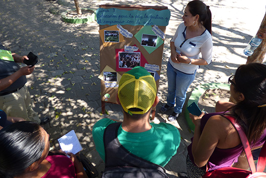 Se muestra una mujer con un cartel exponiendolo frente a varias personas, en una zona rural.